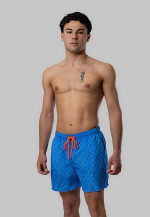 Giovane uomo in piedi, senza maglietta, indossa pantaloncini da bagno blu a fantasia con coulisse arancione e un tatuaggio verticale sul petto con caratteri coreani.