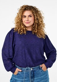 Violeta mežģīņu blūze ar teksturētām dimanta rakstām, garām ballona piedurknēm un apaļu kakla izgriezumu, saskaņota ar augsto vidukli gaišām džinsu biksēm.