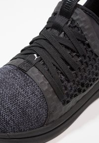 Gros plan d'une basket noire avec un bout en tissu tricoté, des empiècements latéraux en mesh et des lacets noirs croisés sur une languette rembourrée.
