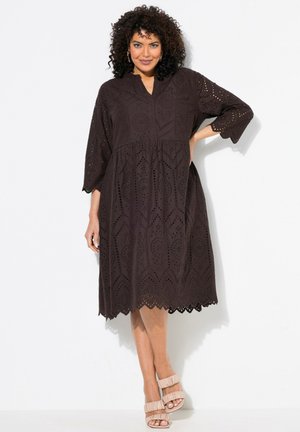 Braunes Augenlet-Kleid mit Dreiviertelärmel, gewelltem Saum und floral gemusterten Ausschnitten. Kombiniert mit hellen, hohen Sandalen.