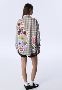 Donna in piedi, che indossa una camicia a quadri verde e rosa con grandi stampe floreali colorate sulla schiena e sulle maniche, pantaloni corti neri, calzettoni bianchi e scarpe nere.