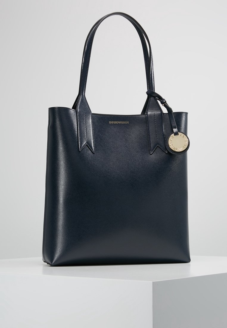 Marineblaue Ledertasche mit strukturierter Oberfläche, zwei Griffen und einem dekorativen Metallanhänger. Die Griffe sind mit eckigen Ausschnitten gestaltet.