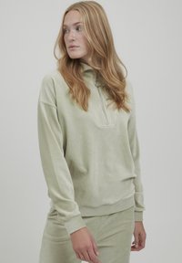 Ljusgrön zip-up tröja i mjukt, texturerat tyg med långa ärmar. Har en liten krage och synliga sömnadsdetaljer.