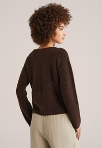 Sieviete ar cirtainiem matiem, valkājot tumši brūnu pūkainu džemperi un gaiši bēšas bikses, stāv neitrālā fonā, skatiens uz sāniem.