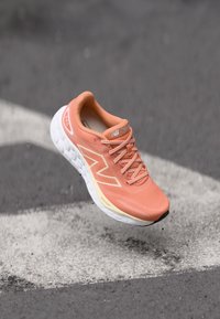 Baskets de course orange clair avec un tige en mesh, semelle intermédiaire blanche et lacets texturés. Présente un logo "N" bien en évidence sur le côté.