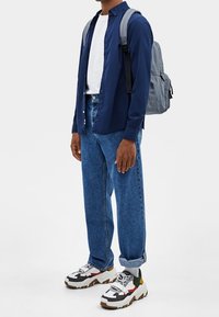 Granatowa koszula na guziki, biała koszulka, luźne niebieskie dżinsy, kolorowe masywne trampki oraz szary plecak z paskami. Swobodny strój.