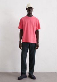 Korte mouw, crew-neck t-shirt van felroze katoen, gecombineerd met donkergroene broek en zwarte instappers, afgemaakt met een lichtbeige hoed.