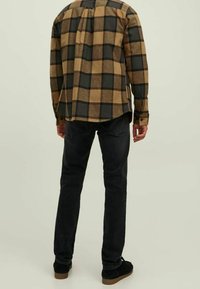 Uomo che indossa una camicia a maniche lunghe a scacchi marroni e neri, jeans neri e scarpe nere, in piedi con la schiena rivolta verso la fotocamera.