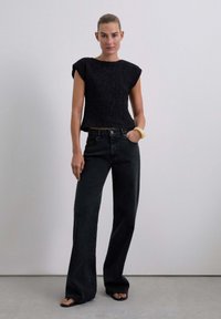 Top corto nero testurizzato con maniche corte, abbinato a jeans neri a vita alta e a gamba larga. Il modello indossa un braccialetto chiaro e sandali.