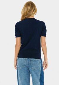 Kvinna med långt blont hår som bär en marinblå kortärmad tröja och tvåfärgade jeans, står vänd bort mot en vit bakgrund.