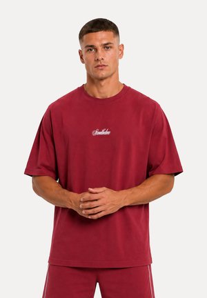 Uomo che indossa una maglietta rossa oversize abbinata e pantaloncini, in piedi con le mani giunte davanti, su uno sfondo chiaro e uniforme.