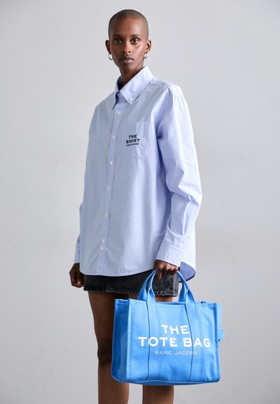 Sac tote bleu avec texte blanc, doté de deux poignées et d'une forme rectangulaire. Porté avec une chemise à rayures bleu clair.