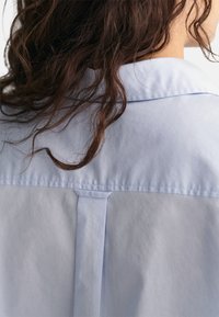 Chemise col bleu clair en tissu lisse et texturé. Présente un empiècement et une couture verticale au dos, mettant en valeur des cheveux longs.