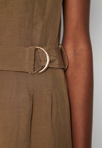 Brun tygklänning med ett brett bälte som har en dubbel guldtonad metallspänne, visad på en persons midja och arm i närbild.