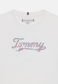 Biała bawełniana koszulka z okrągłym dekoltem, z kolorowym, cekinowym logo "Tommy" z przodu. Metka pokazuje nazwę marki i rozmiar.