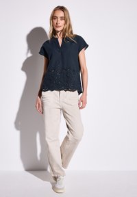 Blusa blu navy a maniche corte con scollatura a V e decorativi fori a occhio sull'orlo, abbinata a pantaloni beige larghi e sneakers chiare.