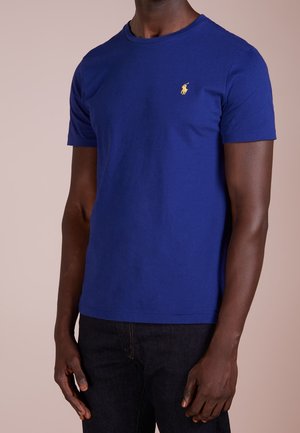 Uomo che indossa una maglietta aderente a maniche corte di colore blu reale con un piccolo logo giallo sul petto a sinistra e jeans scuri.