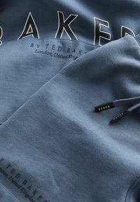 Modrá bavlněná mikina s kapucí s velkými bílými písmeny "BAKER" na přední straně a malou logo nášivkou na lemu. Měkká textura a ležérní design.