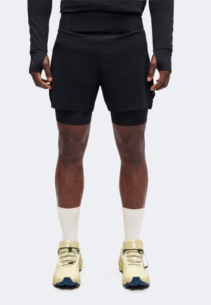 Athlète portant un short noir superposé, un haut noir à manches longues, des chaussettes blanches courtes et des chaussures de trail couleur crème, debout sur un fond clair.