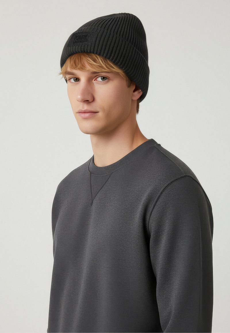 Schwarze, gerippte Mütze aus Strickmaterial mit strukturierter Oberfläche und einem Logo-Patch auf der Vorderseite, getragen zu einem dunkelgrauen Rundhals-Sweatshirt.