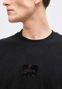 Νεαρό άτομο με μαύρο πουλόβερ χωρίς γιακά που έχει ένα λαμπερό μαύρο τετράγωνο πατς με την επιγραφή "HUGO" στο στήθος, κοιτάζοντας πλάγια.