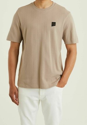 Beżowy T-shirt z krótkim rękawem z teksturowanymi ukośnymi paskami i małą czarną łatką z logo na piersi; noszony z białymi spodniami.