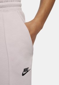 Pantalones deportivos de color rosa claro hechos de tela suave, con cintura elástica y bolsillo lateral, con un logotipo negro de Nike en la parte inferior izquierda.