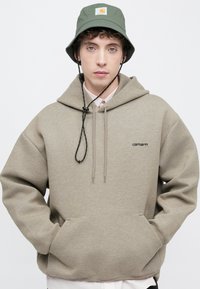Sudadera con capucha de color marrón claro con cordones y bolsillo frontal; presenta el logo negro "carhartt" en el pecho. Combinada con un sombrero tipo bucket verde.