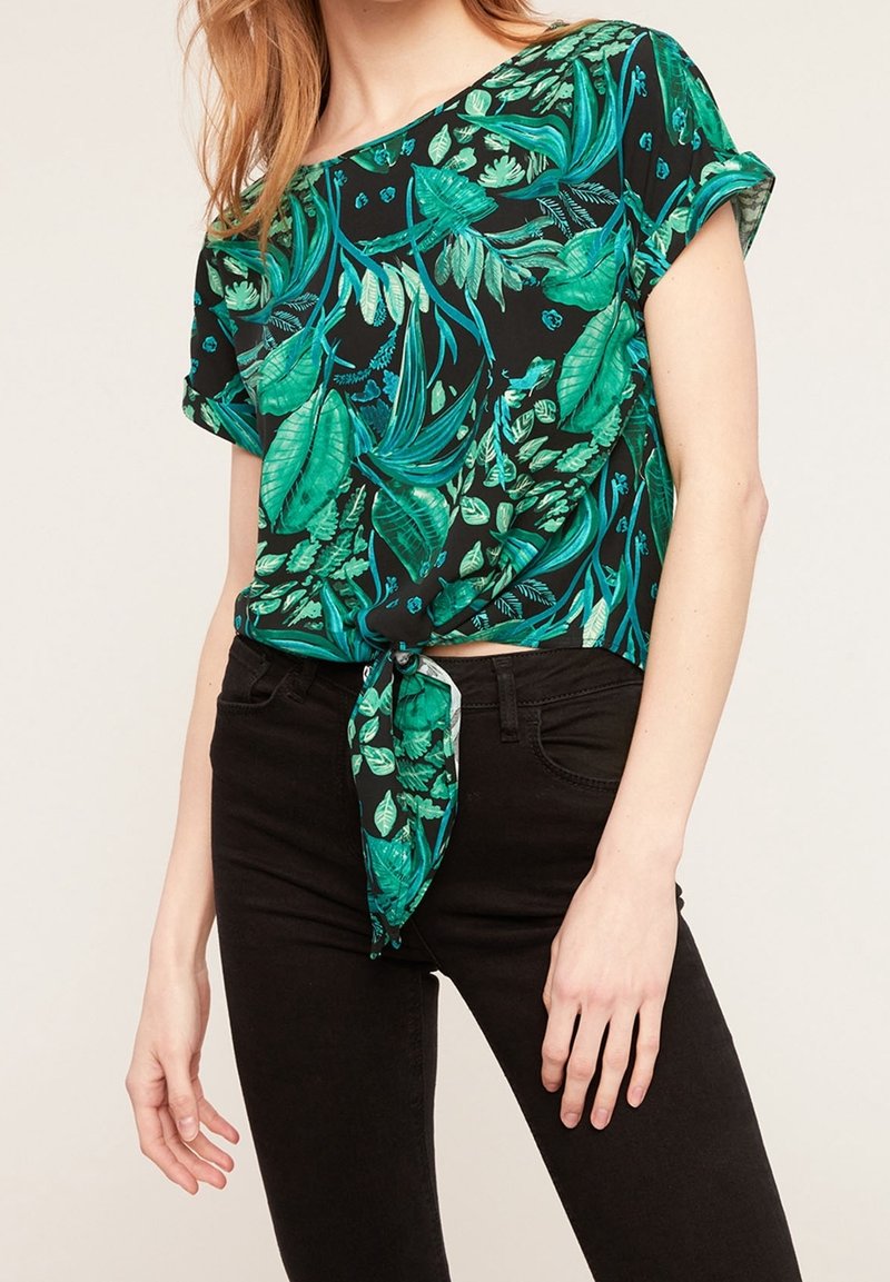Femme portant un jean noir et un haut à manches courtes noir avec un imprimé de feuilles tropicales vertes, noué devant à la taille.