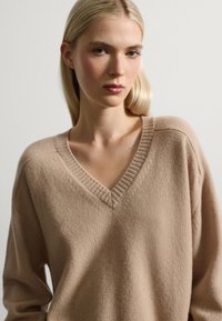 Beige V-hals trui van zacht materiaal, met geribbelde accenten bij de halslijn en een relaxte pasvorm in een lichte textuur.