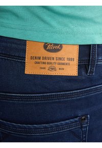 Denim blu scuro con un'etichetta in pelle marrone che riporta "Petrol" e "DENIM DRIVEN SINCE 1989", con cuciture doppie e dettagli delle tasche.