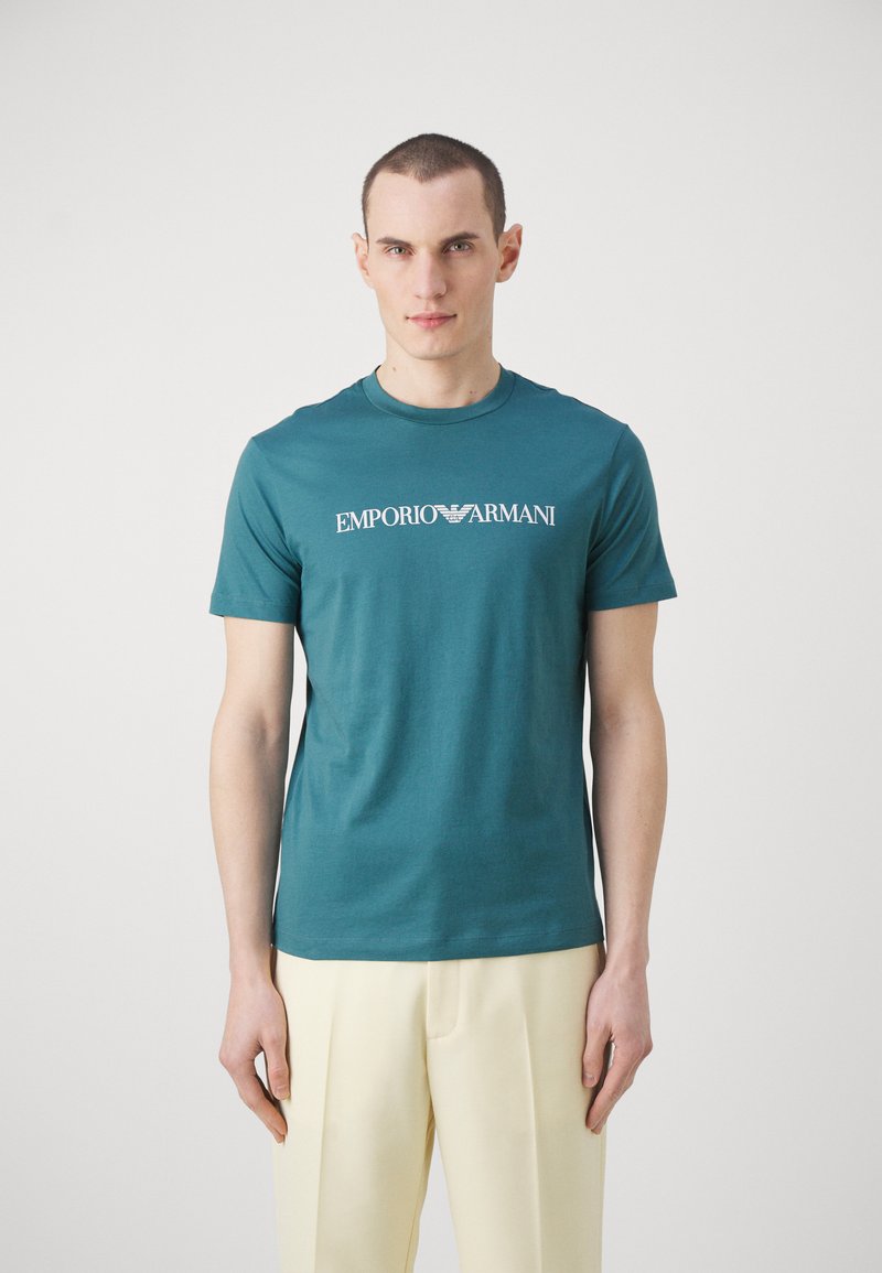 Mėlynos spalvos medvilninė marškinė su apvalia iškirpte ir trumpomis rankovėmis, puoštais balta "Emporio Armani" logotipu per krūtinę. Lygi tekstūra.