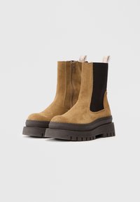 Botas de tobillo de ante marrón con cremallera lateral, panel elástico negro y suela de goma gruesa con estrías. Tablero en la parte trasera para facilitar el calzado.