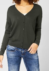 Tmavě zelený svetr s výstřihem do V, dlouhými rukávy a zapínáním na knoflíky. Vyroben z měkké tkaniny. V kombinaci s modrými džíny s dekorativním šitím.