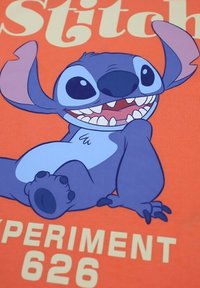 Le personnage de dessin animé Stitch, représenté en bleu avec de grandes oreilles et de grands yeux, sourit sur un fond orange avec le texte "EXPÉRIENCE 626".