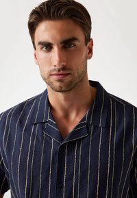 Camisa de rayas en tonos navy, hecha de lino, con líneas verticales en blanco y verde. Presenta mangas cortas y un cuello con muesca, adornado con botones negros.