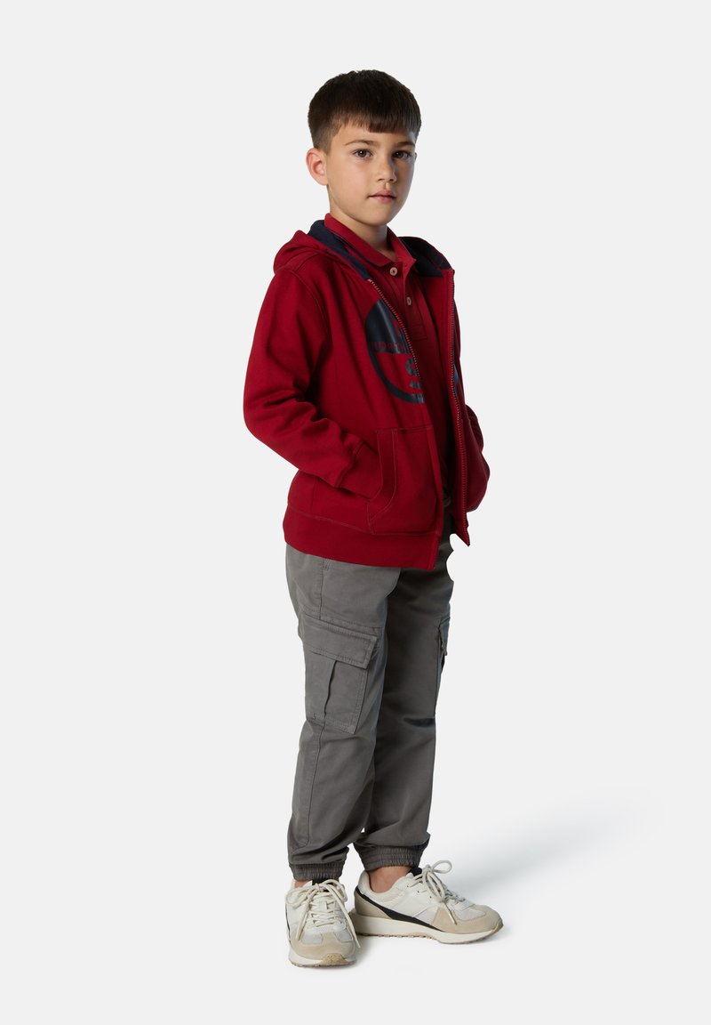Sudadera roja con cremallera y capucha forrada de azul marino, pantalones cargo grises y zapatillas blancas. La sudadera presenta un gráfico circular en el pecho.