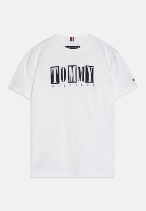 Hvid T-shirt med korte ærmer og sort-hvide blokbogstaver, der staver "TOMMY" med mindre tekst "HILFIGER" under logoet.