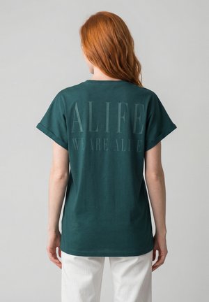 Person mit langen roten Haaren, von hinten gesehen, trägt ein dunkelgrünes T-Shirt mit dem Schriftzug "ALIFE WE ARE ALIFE" auf dem Rücken und weiße Hosen.