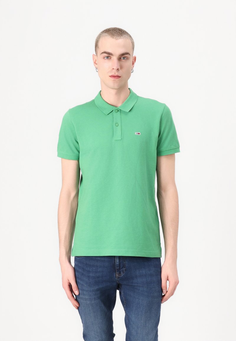 Grønn polo-skjorte laget av bomull, med en klassisk krage og korte ermer, og et lite logo-innslag på venstre bryst. Båret med blå jeans.