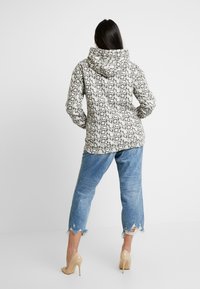 Mikina s kapucí v béžovo-černém geometrickém vzoru, doplněná modrými džínami s potrhaným vzhledem a průhlednými vysokými podpatky.