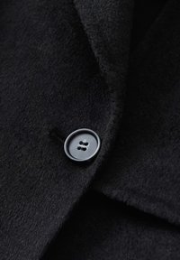 Cappotto di lana nero caratterizzato da una consistenza morbida, colletto a punta e un bottone grigio rotondo con due fori, posizionato sul davanti.
