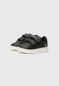 Sapatilhas pretas em pele com duas tiras de velcro, sola branca, detalhes perfurados nas laterais e um logótipo na tira e na lateral.