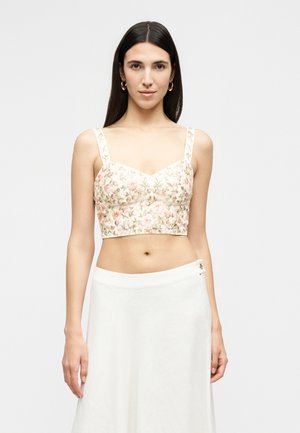 Γυναίκα με μακριά σκούρα μαλλιά φοράει μια κροπ τοπ με κεντήματα λουλουδιών και λευκή φούστα με ψηλή μέση, στέκεται μπροστά σε ένα απλό ανοιχτό φόντο.