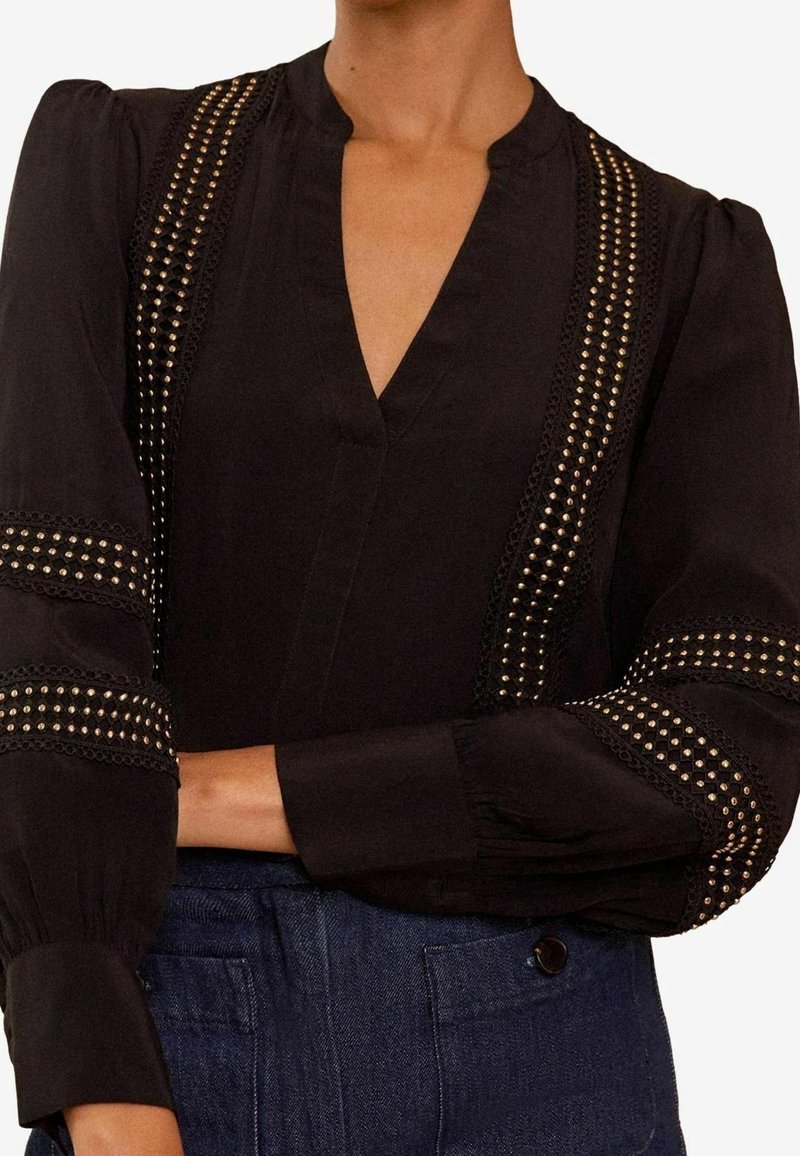 Blouse noire avec détails en dentelle cloutée verticale et sur les manches, manches longues bouffantes, et décolleté en V, associée à un pantalon bleu foncé taille haute.