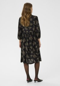 Rochie neagră cu flori, cu tiv până la genunchi, croi lejer și mâneci bufante. Prezintă motive florale închise și este purtată cu tocuri negre.