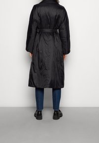 Černá péřová bunda s opaskem v oblasti pasu, dlouhými rukávy a hladkým, lesklým povrchem. Oblečeno s modrými džíny a černými robustními botami.