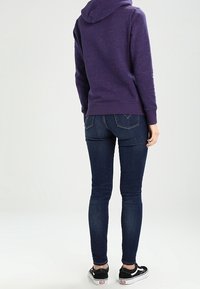 Sudadera púrpura con acabado texturizado, combinada con jeans ajustados de color azul oscuro y zapatillas negras. La sudadera cuenta con capucha y puños acanalados.