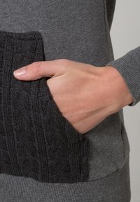 Maglione grigio con una tasca lavorata a maglia in grigio scuro. Mano inserita nella tasca, che evidenzia il contrasto tra i materiali.