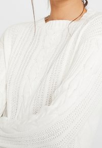 Woman wearing a white cable-knit sweater with braided and open-knit patterns, showing upper torso and part of neck and hair strands.
